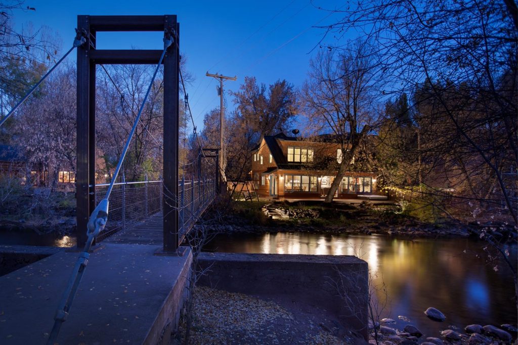 A suspension bridge leads across a small river to a warmly-lit home at night